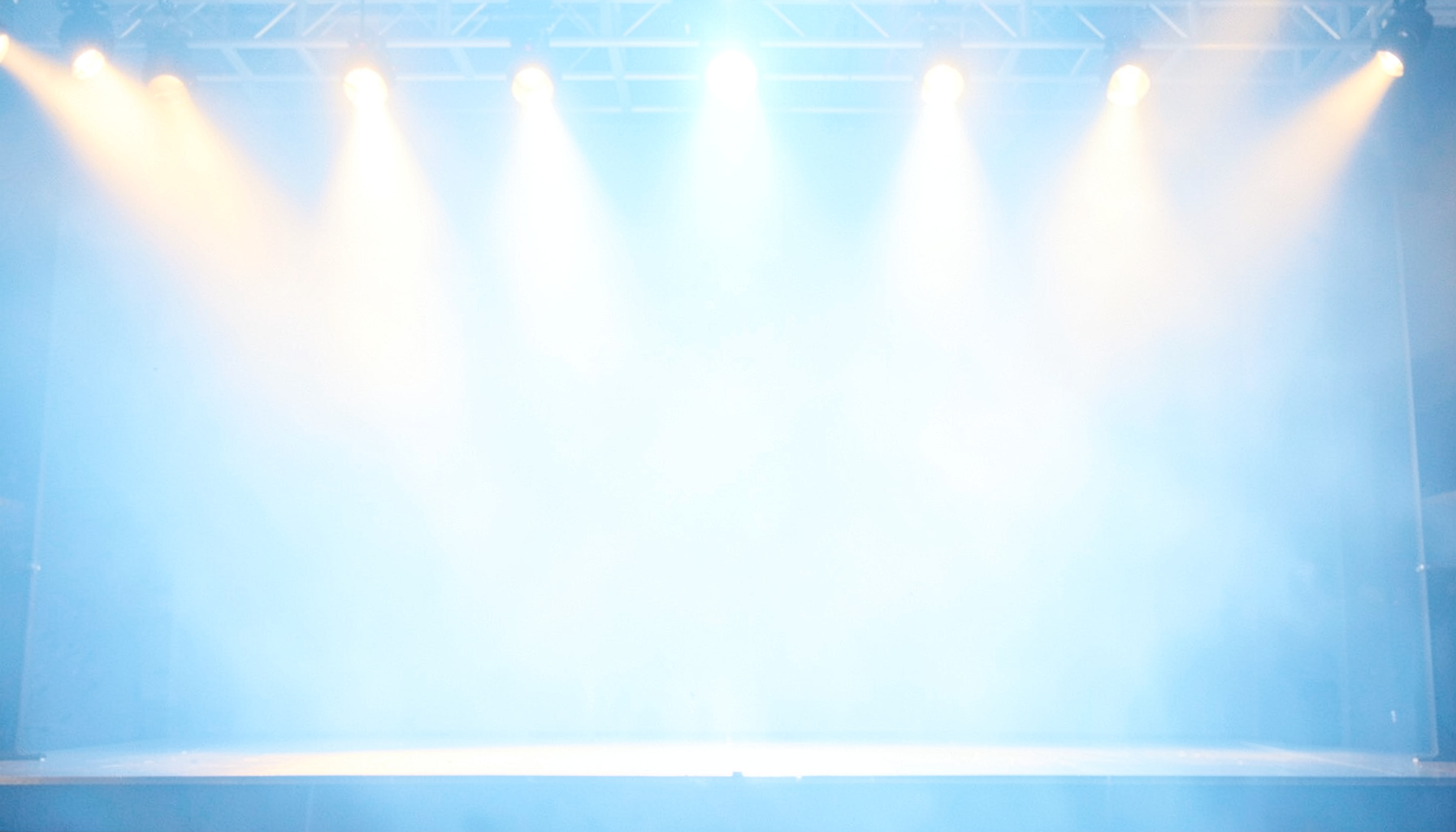 A bright, airy high-key photograph of a softly lit stage with warm amber spotlights against pale blue haze, evoking a live performance atmosphere