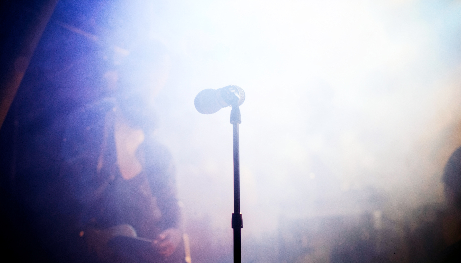 A warm stage-lit performance scene with deep indigo and amber tones, a microphone on a stand silhouetted against soft haze