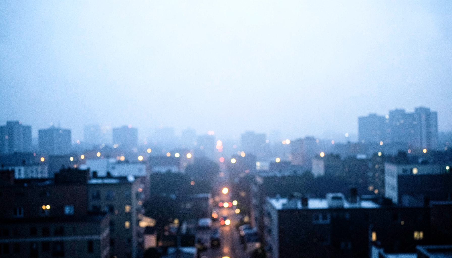 A soft, atmospheric nighttime cityscape in deep navy and charcoal tones, with distant warm amber streetlights glowing through haze